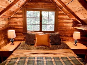 A cozy attic bedroom with a large window, a bed with a plaid blanket, two bedside lamps, and a pinecone decoration.