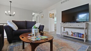 A living room with a dark-colored sofa, a coffee table with magazines, a television on a stand, and a patterned rug. - Wheels Trails and Tails Inn - Fully Fenced! 2x King 2x Twin Pet Friendly (Boonville)