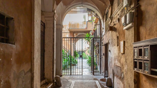 A narrow alleyway with a cobblestone path leading to a gated entrance, flanked by old buildings with balconies and potted plants. - Al Cortile del Moro Suite (Roma)