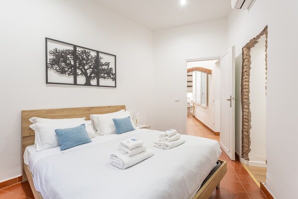 A hotel room with a wooden bed, white bedding, blue pillows, and a framed artwork of a tree. - 2 BDR Navona Quintessential Charm (Roma)