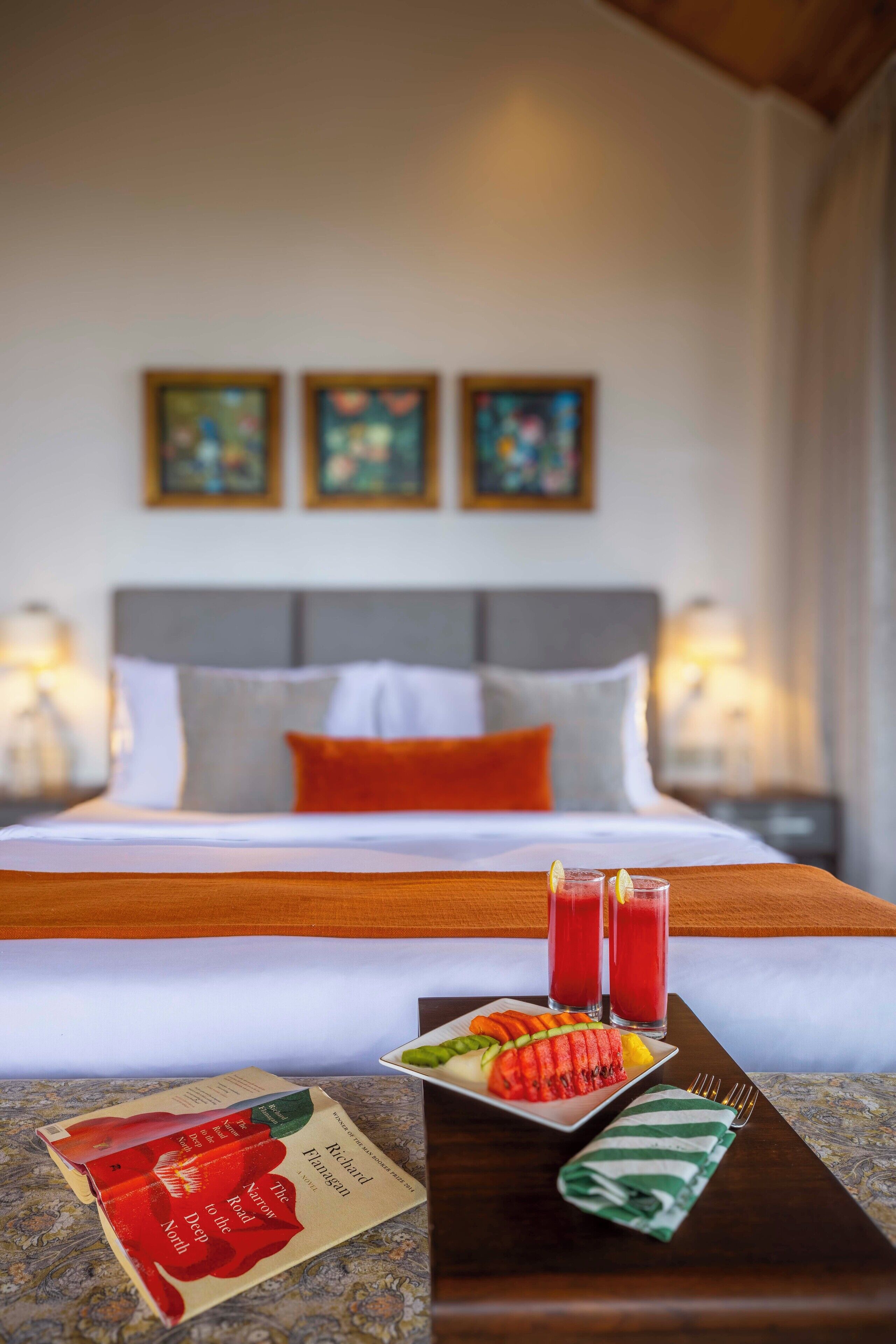 A neatly made bed with a white and orange pillow arrangement, a bedside table with a tray of food and drinks, and a book titled "The Narrow to the Deep North" by Richard Flavin.