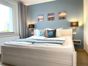 Een bed met een wit hoofdeinde, een raam met gordijnen en wandlampen.