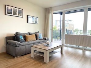 Een moderne woonkamer met een grijze bank, een houten salontafel en een groot raam met uitzicht op gebouwen en een balkon.