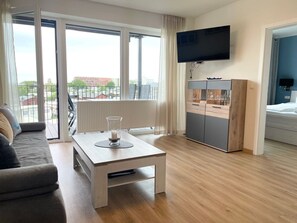 Ein modernes Wohnzimmer mit einer Couch, einem Couchtisch, einem an der Wand montierten Fernseher und Blick durch große Fenster.
