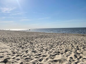 Ein sandiger Strand mit Fußspuren, die zum Wasser führen, ein klarer Himmel und ein Pier in der Ferne.