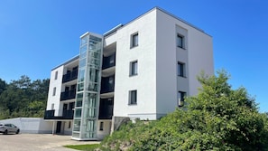 Ein modernes, mehrstöckiges Gebäude mit einem Glasturm für den Aufzug, einem geparkten Auto davor und üppiger Bepflanzung ringsum.