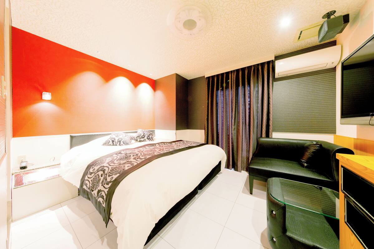 A modern hotel room with a large bed, a desk, a flat-screen TV, and a seating area with a sofa and curtains.