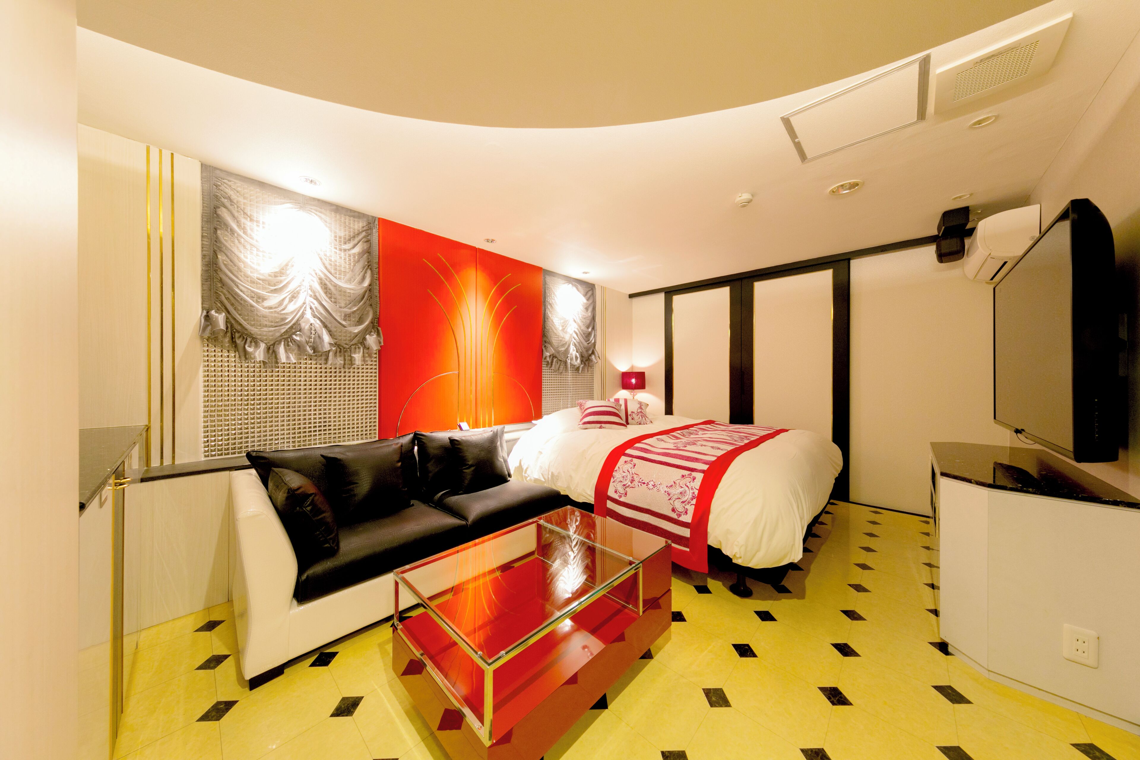 A modern hotel room with a curved ceiling, a glass coffee table, a black sofa, a bed with a red and white bedspread, and a flat-screen TV.
