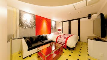 A modern hotel room with a curved ceiling, a glass coffee table, a black sofa, a bed with a red and white bedspread, and a flat-screen TV.