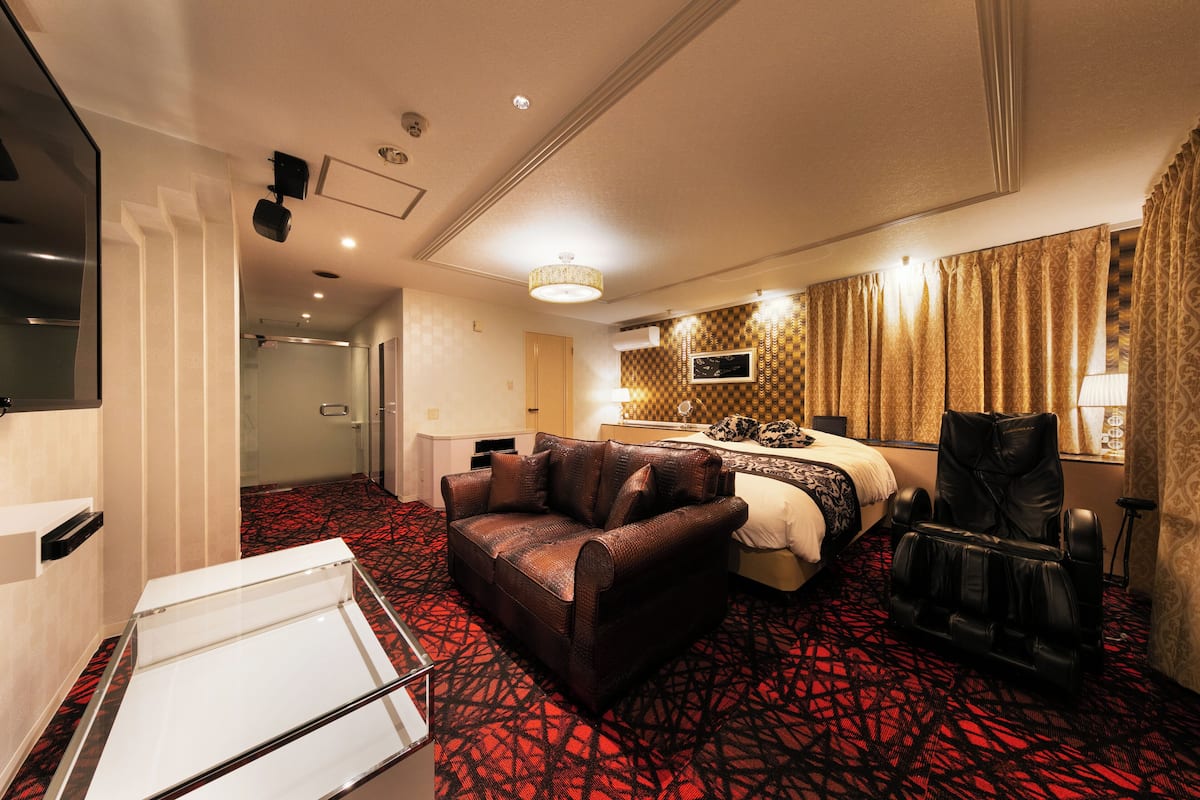 A hotel room with a red patterned carpet, a brown leather sofa, a massage chair, a bed with a patterned headboard, and a television on the wall.