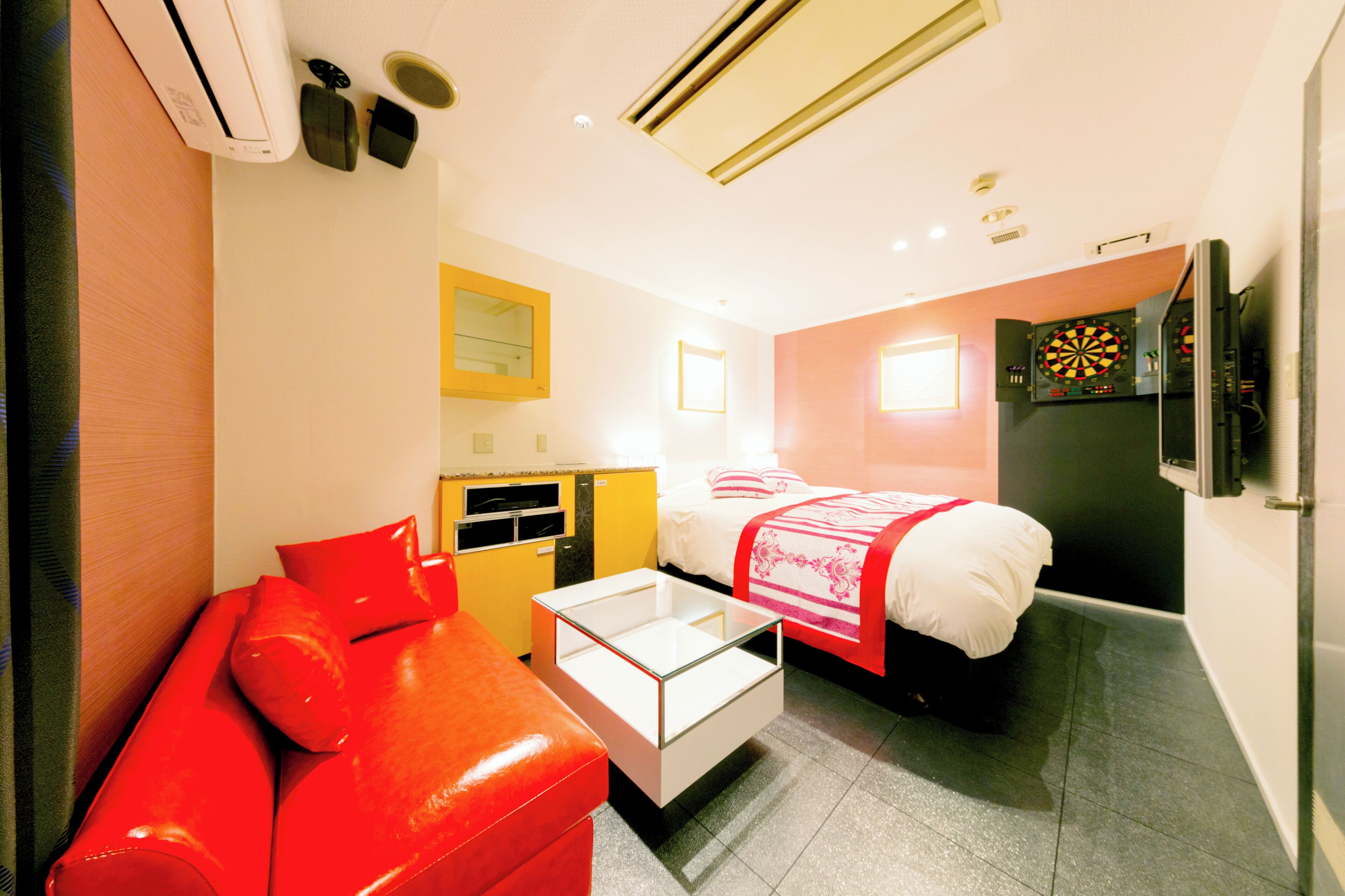 A compact hotel room with a bed, a red sofa, a small table, and a dartboard on the wall.