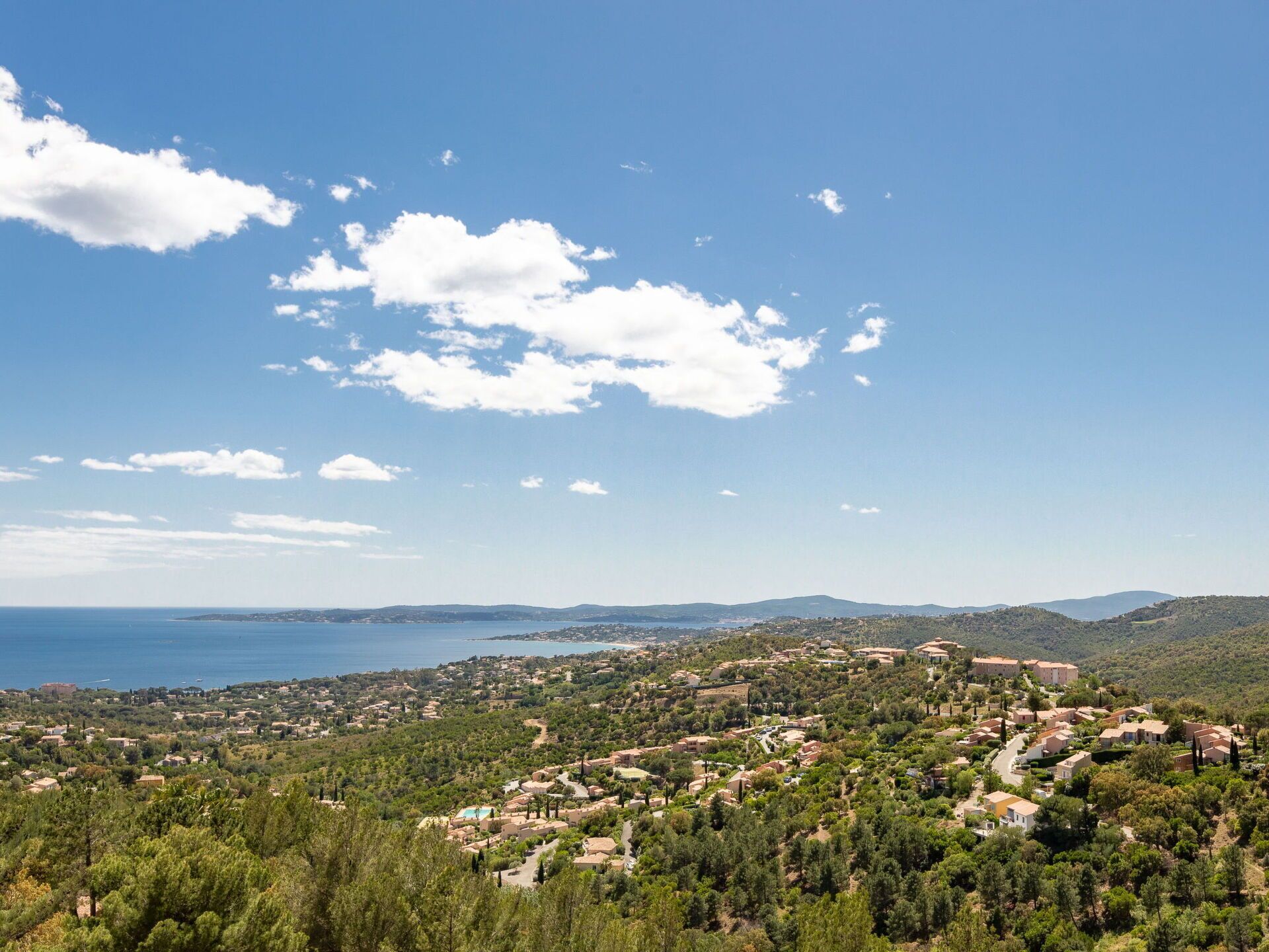 A coastal town with a mix of residential buildings and greenery, a clear sky, and a view of the sea.