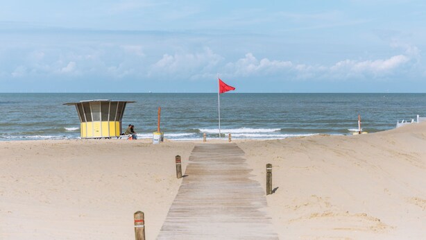A wooden boardwalk leading to a lifeguard station with a red flag on a beach.