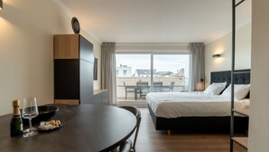 A hotel room with a bed, a dining table with chairs, a small balcony, and a view of the cityscape.