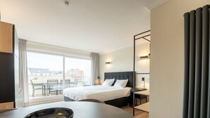A hotel room with a bed, a desk, a chair, a dining table with a bowl and snacks, and a view of the city through large windows.