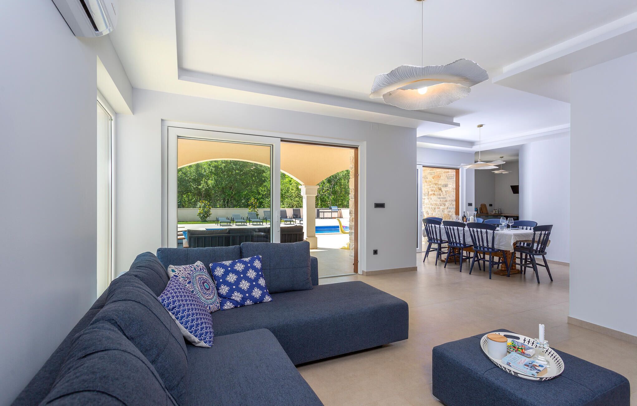Uma sala de estar moderna com um sofá modular cinza, uma área de jantar com mesa e cadeiras, e vista para uma área de piscina externa.