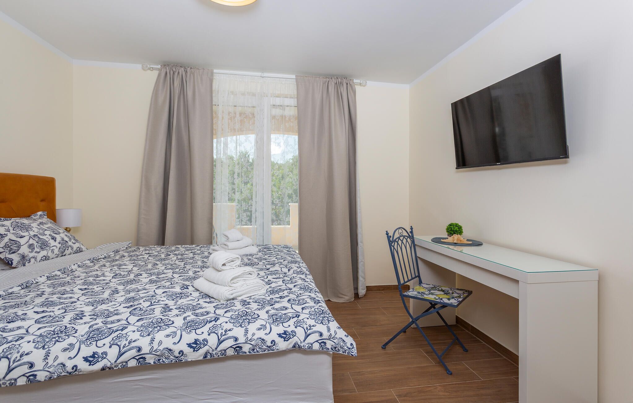 A bedroom with a bed, a desk, a chair, a TV, and a window with curtains.