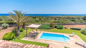 Une piscine agrémentée d’une pergola et de chaises longues, entourée de verdure et offrant une vue sur la mer.