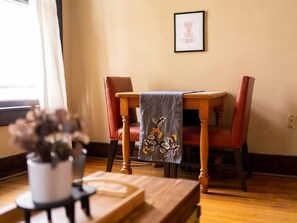 Uma sala de jantar com uma mesa de madeira, duas cadeiras vermelhas, uma frase emoldurada na parede e um vaso com planta no chão.