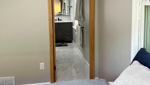 Un dormitorio con cama, ventana con persianas y vista a un baño con espejo, lavamanos y toallero.