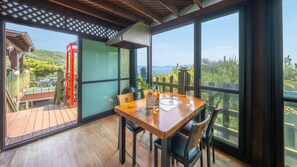 Un comedor con una mesa y sillas de madera, ventanas amplias que ofrecen vistas a una estructura roja y follaje, y una terraza de madera en el exterior.