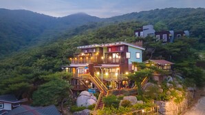 Una casa de varios pisos con un balcón de madera y escaleras, rodeada de abundante vegetación y montañas.