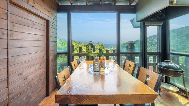 Una mesa de madera con sillas, un jarrón y una botella, frente a amplios ventanales con vistas a un paisaje exuberante.