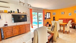 A living room with a TV, wooden cabinet, dining table, chairs, and a view of a pool through large windows. - Villa LOS JAZMINES at Monte Pego (Pego)