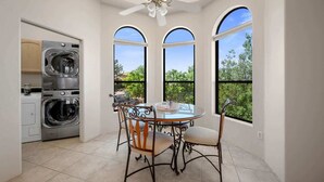 Un lavadero con lavadora y secadora, una zona de comedor con mesa y sillas, y amplias ventanas de arco que ofrecen vistas a la vegetación.