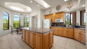Una cocina amplia con armarios de madera, encimera de granito y una isla grande. Hay tres ventanas que brindan luz natural y vistas al exterior.