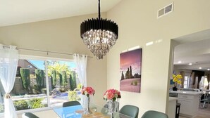 A dining area with a glass table and green chairs, a chandelier, and a view of the outdoors. - Family Paradise near Disneyland w/ Sparkling Pool (Fountain Valley)