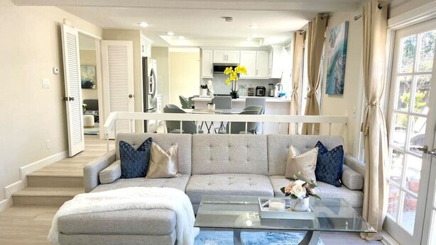 A living room with a sofa, ottoman, and coffee table, a kitchen in the background, and a large window with curtains. - Family Paradise near Disneyland w/ Sparkling Pool (Fountain Valley)