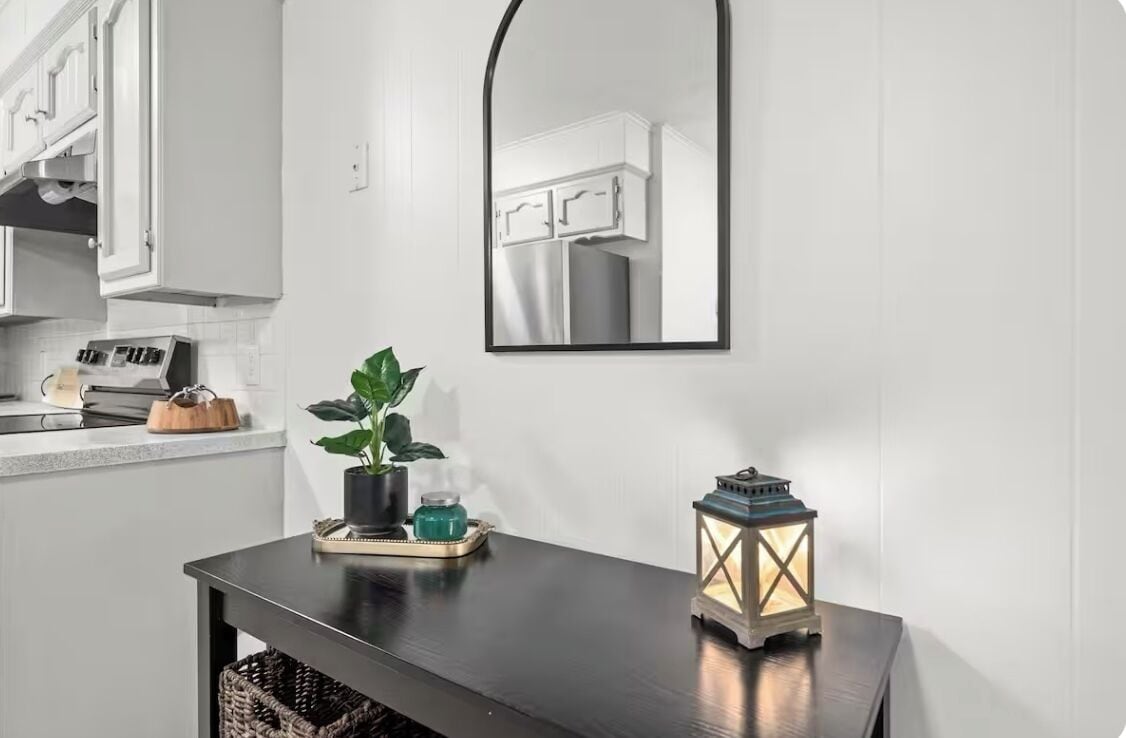 A modern kitchen with white cabinets, a black countertop, a wooden table with a potted plant, a lantern, and a mirror reflecting the cabinets.
