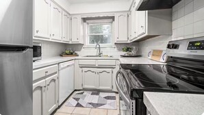 A modern kitchen with white cabinets, a stainless steel refrigerator, a black stove, and a window with blinds.