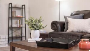 A living room with a dark brown coffee table, a white vase with green plants, a black tray, a red candle, a dark brown sofa with a white pillow, and a bookshelf in the background.