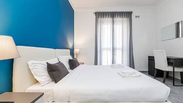A hotel room with a bed, a desk, a chair, a lamp, and a window with curtains.