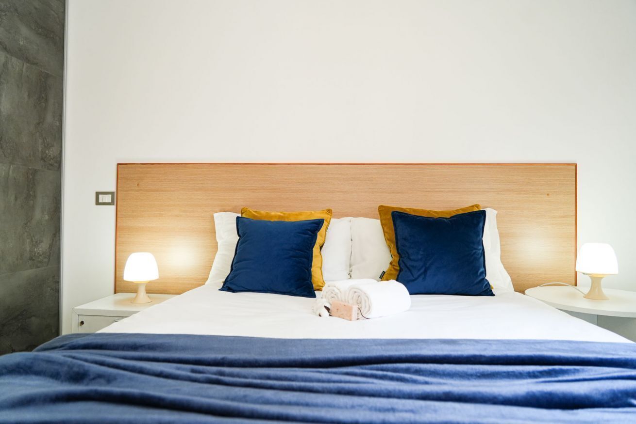 Una cama bien tendida con una colcha azul, dos almohadas en azul y amarillo, y un objeto blanco pequeño sobre la cama.
