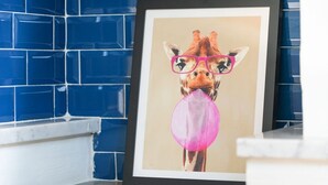 A framed picture of a giraffe wearing pink glasses and holding a pink balloon, set against a blue tiled wall. - Torino Centro, Art House Arsenale (Torino)