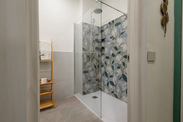 A glass-enclosed shower with a floral tile design, a wooden shelf with towels, and a wall-mounted light fixture.