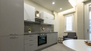 Une cuisine moderne avec des meubles gris, un four encastré et un coin repas comprenant une table et des chaises blanches.