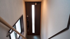 A narrow hallway with a wooden door at the end, a visible emergency exit sign, and a small window to the left. - HANASTAY YANAGI (Osaka)