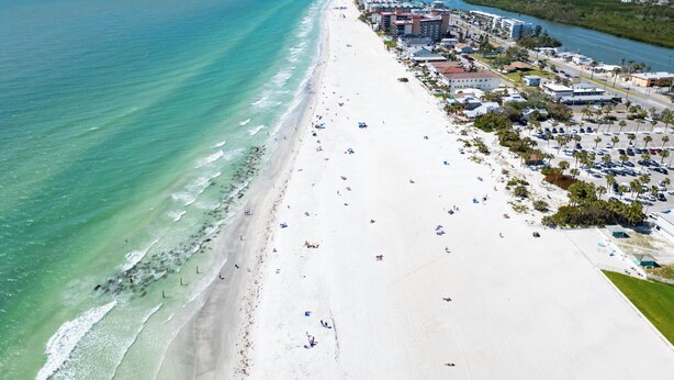 Vista aérea de una playa con una extensa franja de arena blanca, un océano azul cristalino y algunos edificios a lo largo de la orilla.