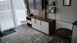 A modern living room with a large flat-screen TV, wooden cabinets, a small round table, a grey sofa, and a balcony with a white railing. - 3-Bed Caravan - Haven Lakeland Leisure Park (Grange-over-Sands)