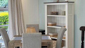 A dining area with a wooden table and four grey upholstered chairs, a white bookshelf with decorative items, and a view of greenery outside.