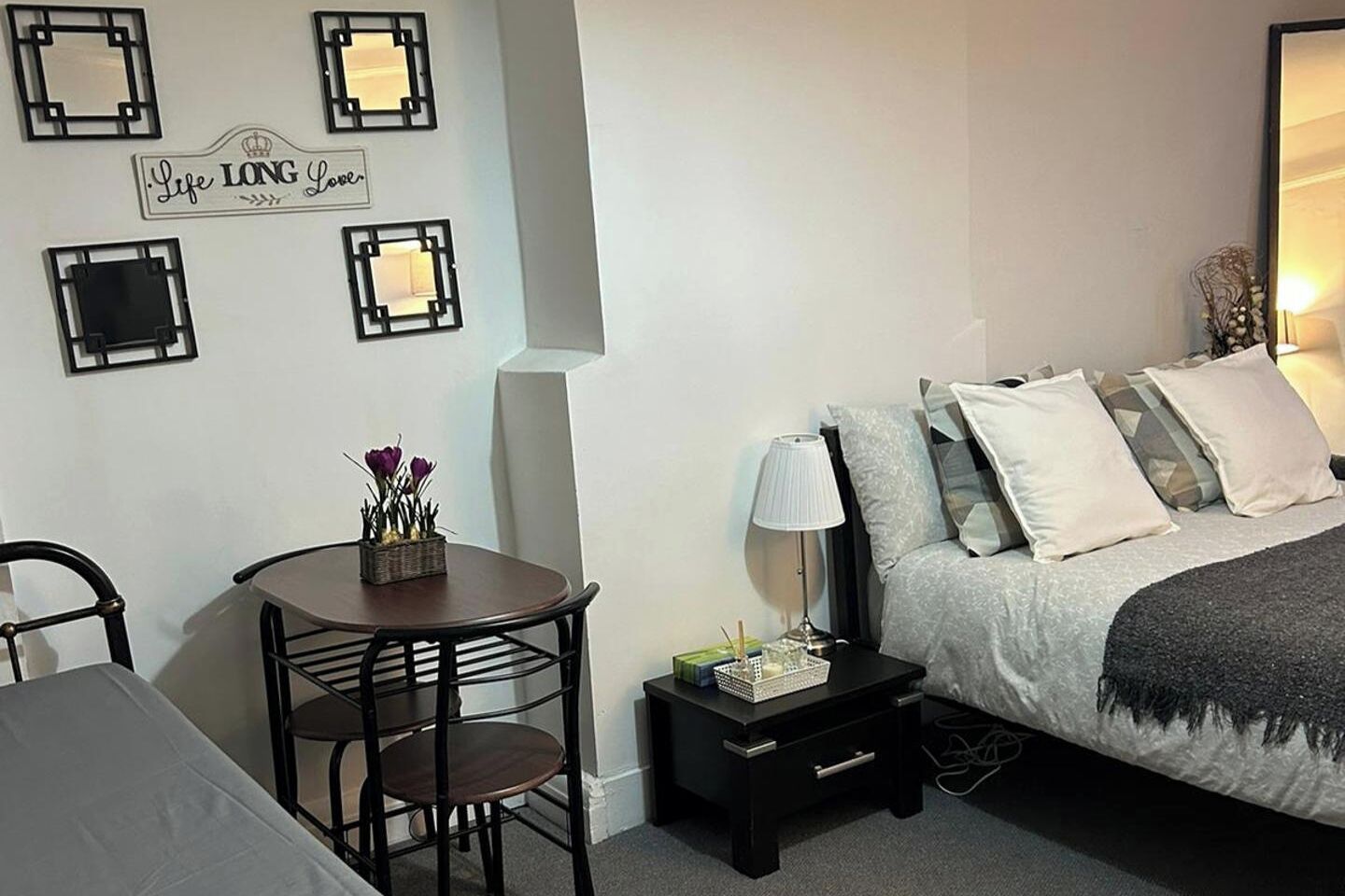 A bedroom with a bed, bedside table, lamp, and a small table with flowers. There are wall decorations and a chair.