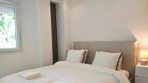 Een net opgemaakt bed met witte lakens en twee opgevouwen handdoeken, een bedlampje en een raam met jaloezieën.