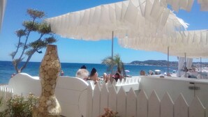 Een terras aan het strand met een witte hek, een decoratief stenen beeld en parasols voor schaduw.
