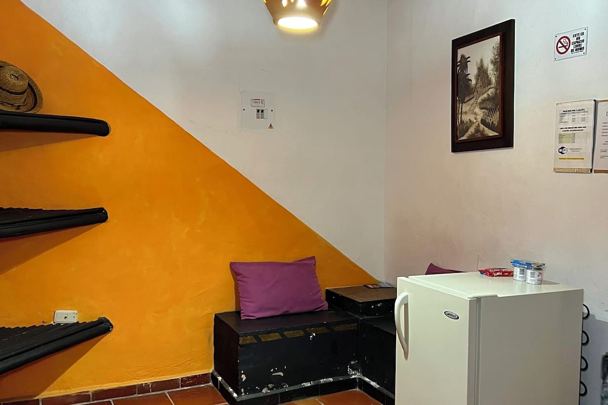 Une pièce avec un réfrigérateur blanc, un coussin violet posé sur une valise noire, un luminaire suspendu et un tableau encadré accroché au mur.