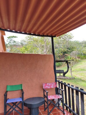 Un espacio de estar al aire libre techado, con dos sillas, una mesa, un banco de madera y vista a un área verde con árboles.