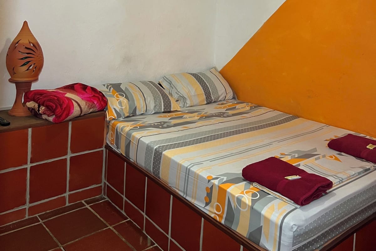 Une petite chambre accueillante, avec un lit simple, une table de chevet, une lampe fixée au mur et un vase décoratif.